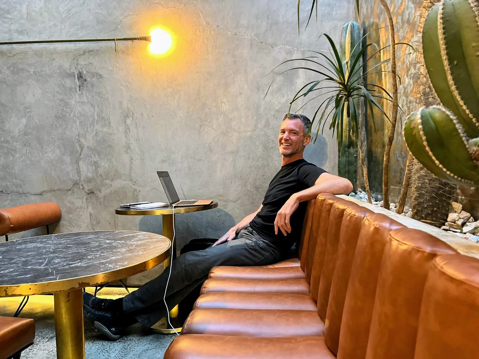 Andrew smiling on a leather banquette with his laptop, in a Mexico City café among cacti