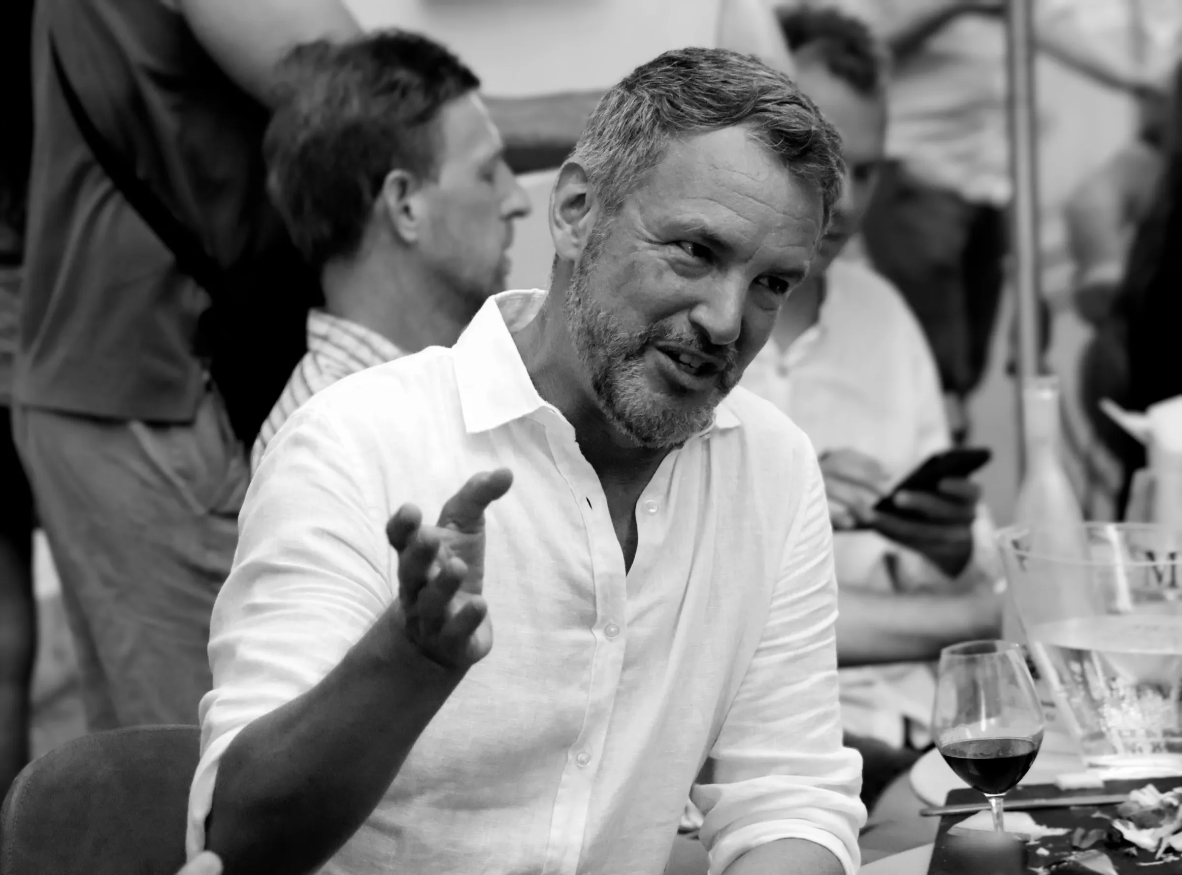 Black-and-white portrait of Andrew at a dinner table, mid-conversation, gesturing with one hand